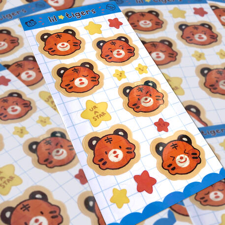 image of multiple 'lil tiger' sticker sheets scattered on top of each other. the sticker sheet shows various facial expressions of a little tiger (a wink face, a tired face, and angry face, a squint face, a happy face, and an aloof face) along with little