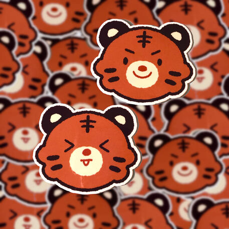 image shows two different 'lil tiger' stickers scattered around. the two designs show different facial expressions of the tiger. One makes a squint face with its tongue out while the other looks more mischievous.